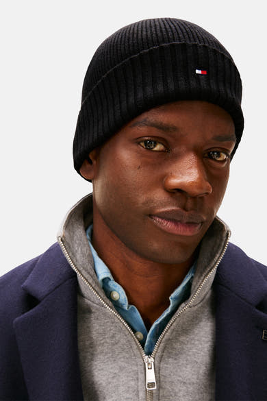 Zwarte gebreide muts van Tommy Jeans, versierd met het merklogo, gedragen met een blauwe jas, een grijze trui en een blauw overhemd.