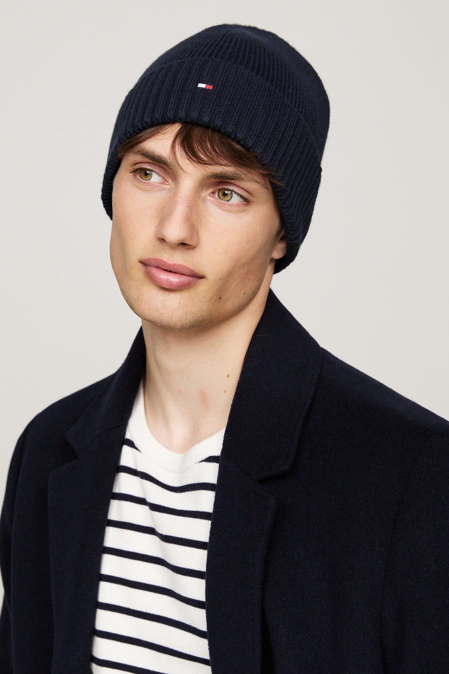 Blauwe gebreide muts van Tommy Jeans, gedragen met een wit en marineblauw gestreepte trui en een marineblauwe jas.