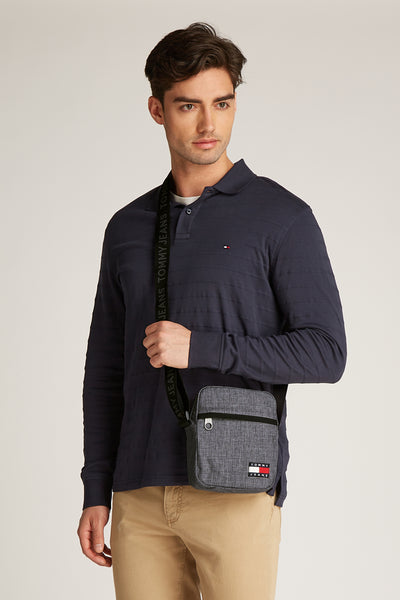 Grijze schoudertas van TOMMY JEANS gedragen door een man, met marineblauw poloshirt en beige broek.
