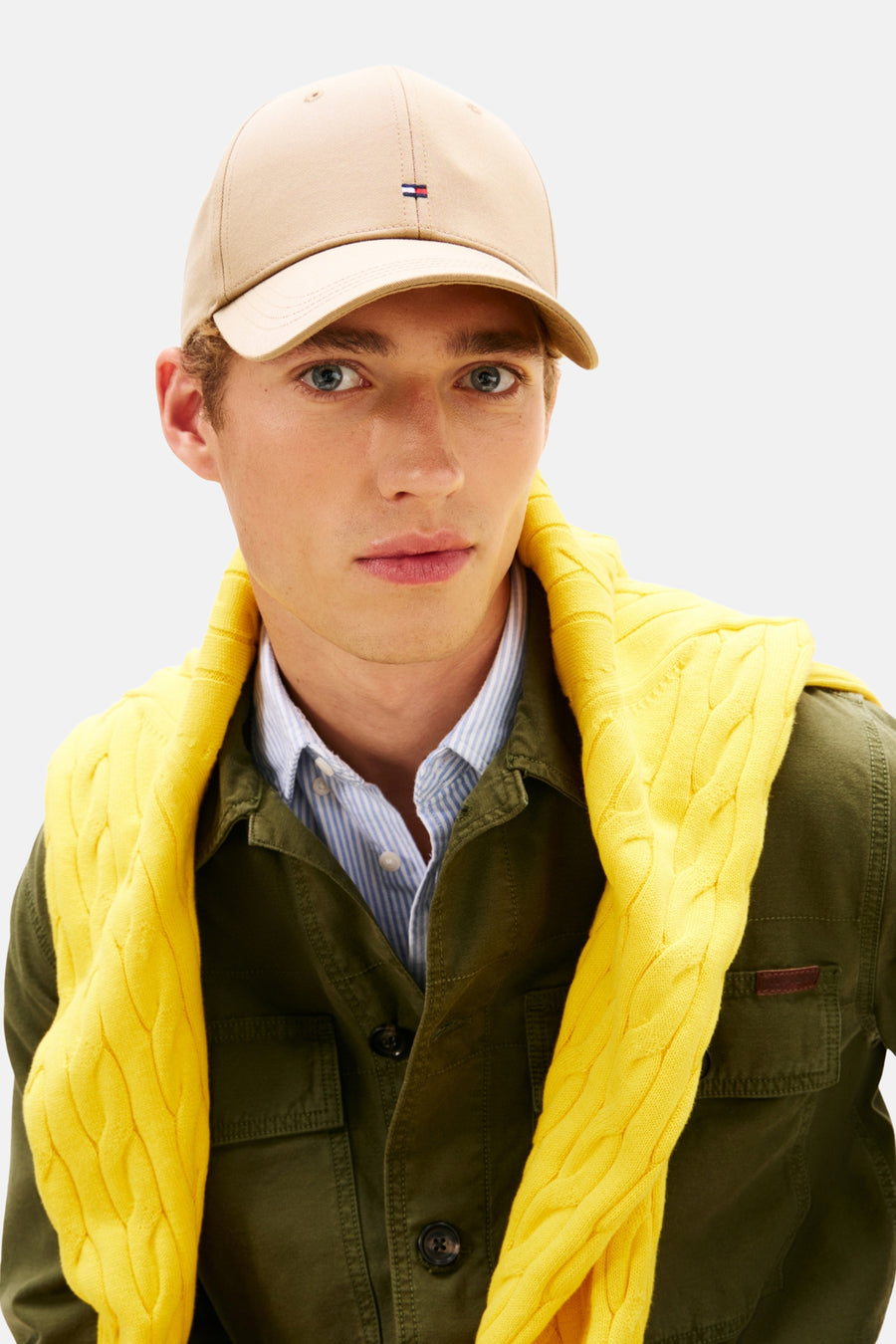 Casquette beige de TOMMY JEANS, avec logo et portée par un homme portant une chemise rayée, une veste et un pull jaune.