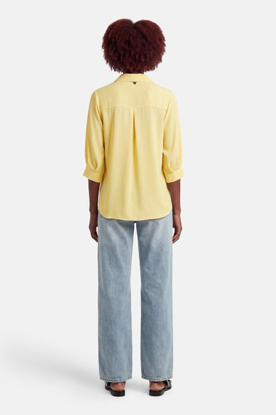 Chemisier jaune à manches courtes par AMELIE ET AMELIE, avec des plis décoratifs au dos et porté avec un jean ample.