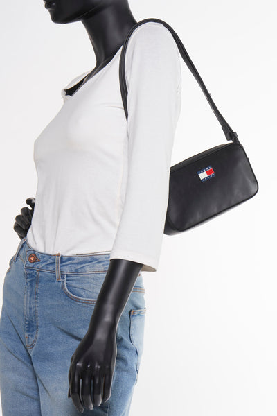 Sac à bandoulière noir de TOMMY JEANS, porté par un mannequin avec un t-shirt blanc et un jean bleu.
