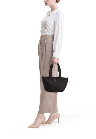Sac à bandoulière noir de Tommy Jeans avec poignées logotypées, porté avec chemisier blanc et pantalon beige.
