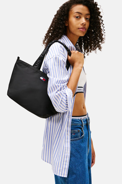 Sac à bandoulière noir de TOMMY JEANS, porté à l'épaule, avec une chemise rayée et un jean taille haute.