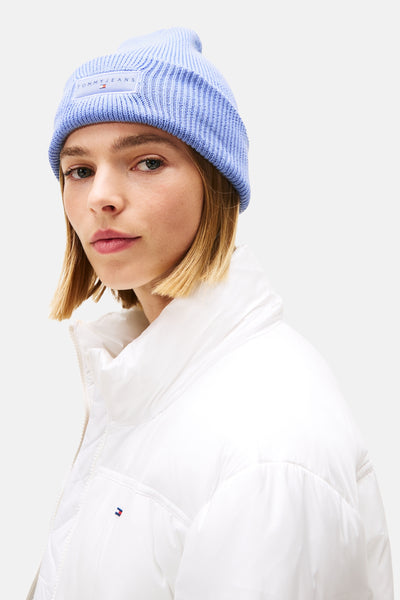 Blauwe gebreide muts van TOMMY JEANS, gedragen met een witte gewatteerde jas met ritssluiting.