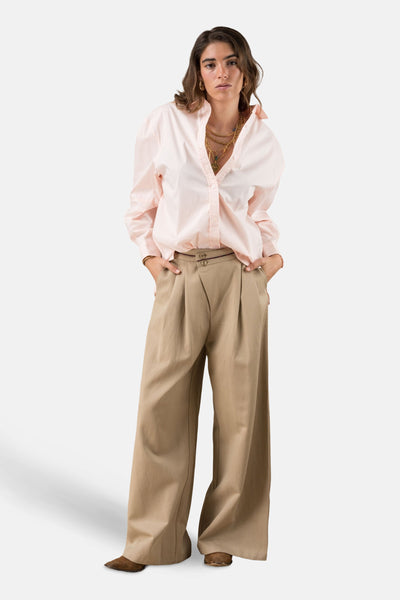 Chino beige ADORE avec plis, porté avec une chemise rose et des colliers dorés.
