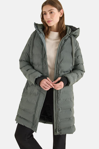 Veste longue verte de CECIL, avec capuche et fermeture éclair sur un pull beige et un pantalon noir.