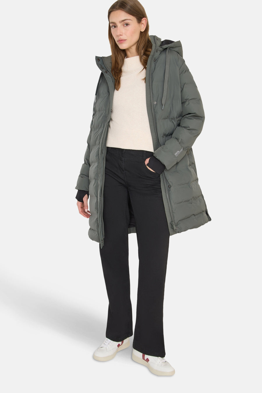 Veste longue vert olive de CECIL, avec capuche et poches, portée avec un pull blanc et un pantalon noir.