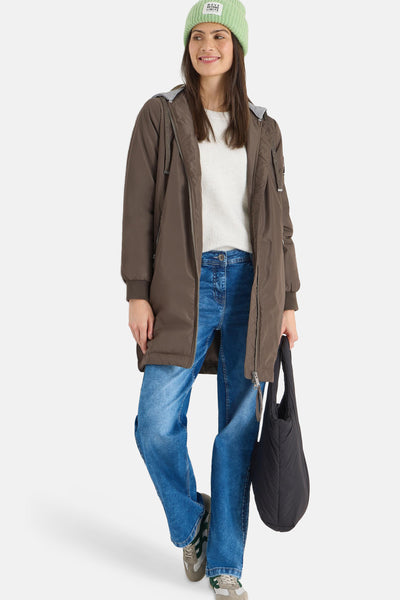 Veste longue brune de CECIL, avec capuche grise et fermeture éclair, portée avec un pull blanc, un jean et un sac noir.