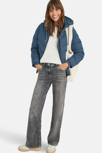 Korte blauwe jas van CECIL, met capuchon en crème fauxbont voering, gedragen met een witte trui en grijze jeans.