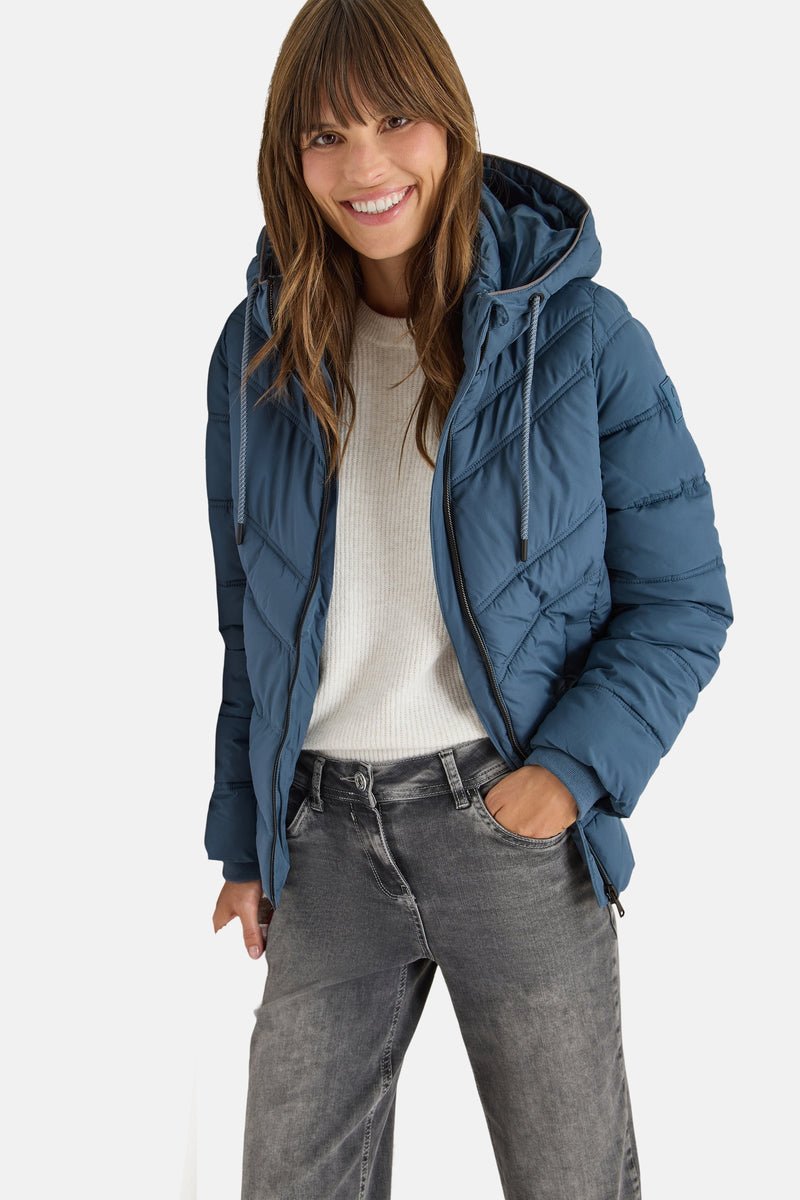 Korte blauwe jas van CECIL, met capuchon en trekkoord en diagonaal doorgestikt patroon, gedragen met een lichte trui en een grijze jeans.