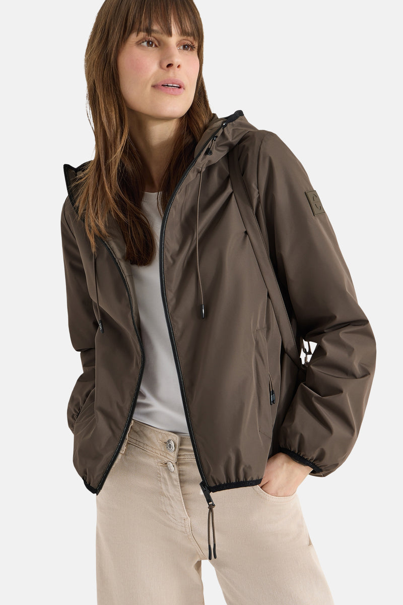 Veste courte brune de CECIL, avec capuche à cordon, portée sur un t-shirt blanc et un pantalon beige.
