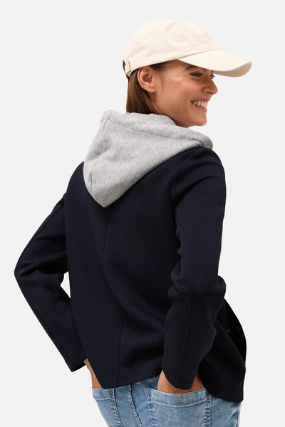 Korte blauwe jas van CECIL, met een grijze capuchon, gedragen met een spijkerbroek en een beige pet.