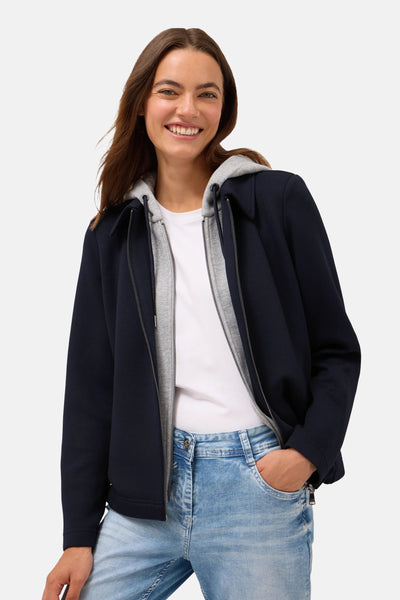 Korte blauwe jas van CECIL met grijze capuchon, gedragen over een wit T-shirt en een lichtblauwe jeans.