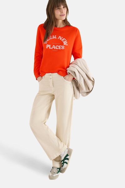 Sweat orange de CECIL avec inscription blanche, porté avec un pantalon beige et des baskets.