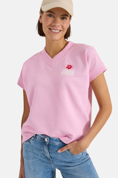 Pink short-sleeve V-neck sweatshirt by Mer&soleil with red coral logo, worn with blue jeans and beige cap, front view.