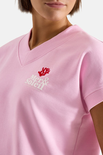 Pink short-sleeve sweatshirt with V-neck and ribbed trim, featuring white "Mer & Soleil" logo with red coral graphic, worn by model in close-up front view.