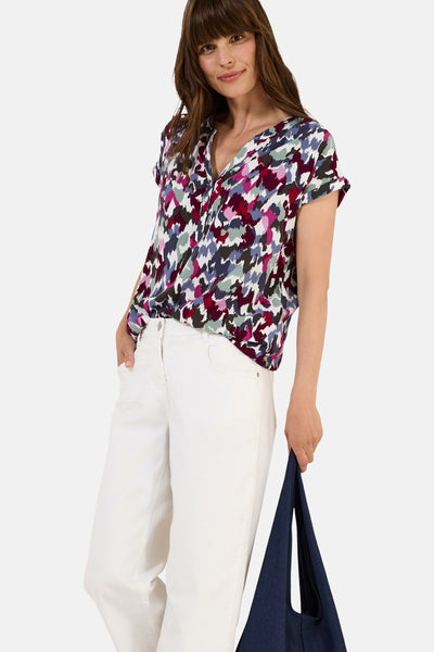 Blouse à manches courtes multicolore de CECIL, à motif abstrait, portée avec un pantalon blanc et un sac bleu marine.
