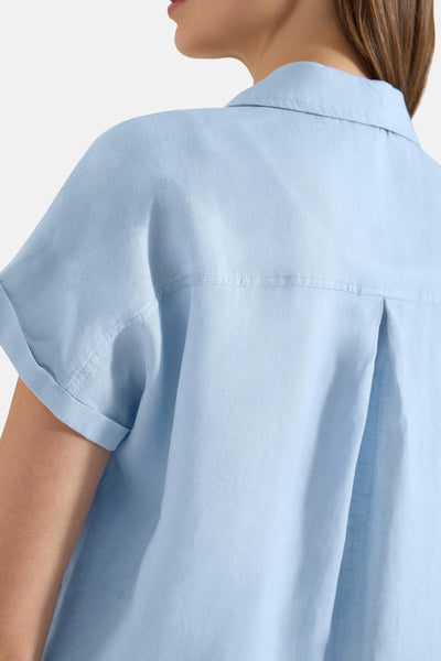 Light blue short-sleeve blouse with collared neckline and pleated back detail, side view of model wearing it.