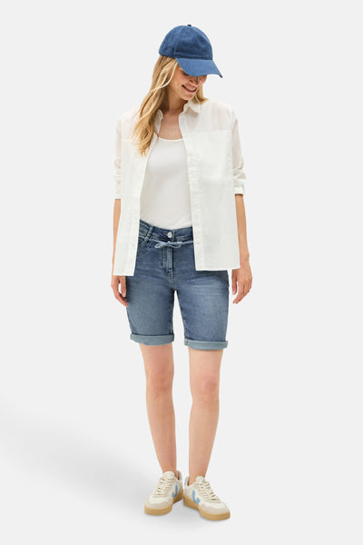 Lichtblauwe denim Bermuda van Cecil, met opgerolde zomen, gedragen met een wit overhemd en hemdje, en een blauwe pet.