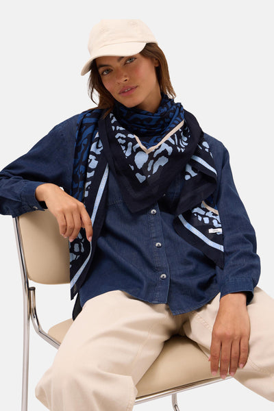 Multikleurige sjaal van CECIL, met een blauw abstract patroon, gedragen met een spijkerblouse en een lichte pet.