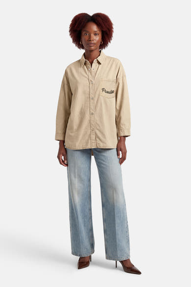 Chemise beige à manches longues par ADORE, avec le mot « Paradise » brodé sur la poche poitrine, portée avec un jean bleu et des escarpins marron.
