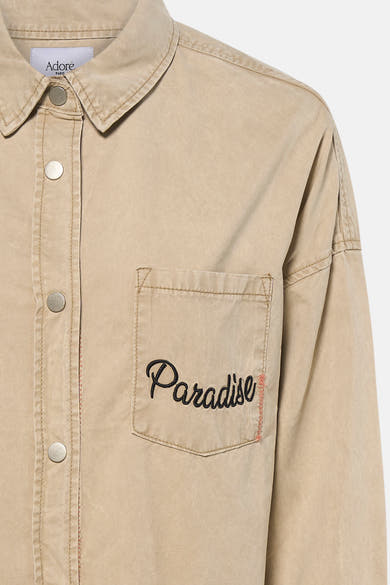 Chemise beige à manches longues par ADORE, avec poche poitrine brodée du mot « Paradise ».
