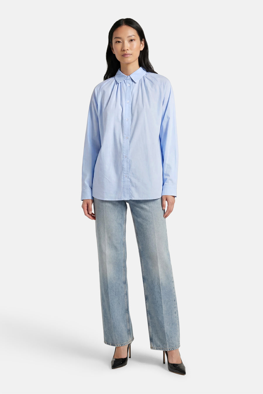 Chemise bleue à manches longues de ADORE, avec un col rond et des plis décoratifs, portée avec un jean et des escarpins noirs.
