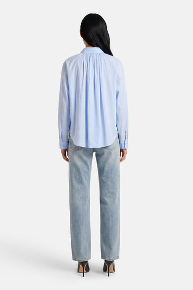 Chemise bleue à manches longues de ADORE, avec détails plissés au dos, portée avec un jean et des talons noirs.
