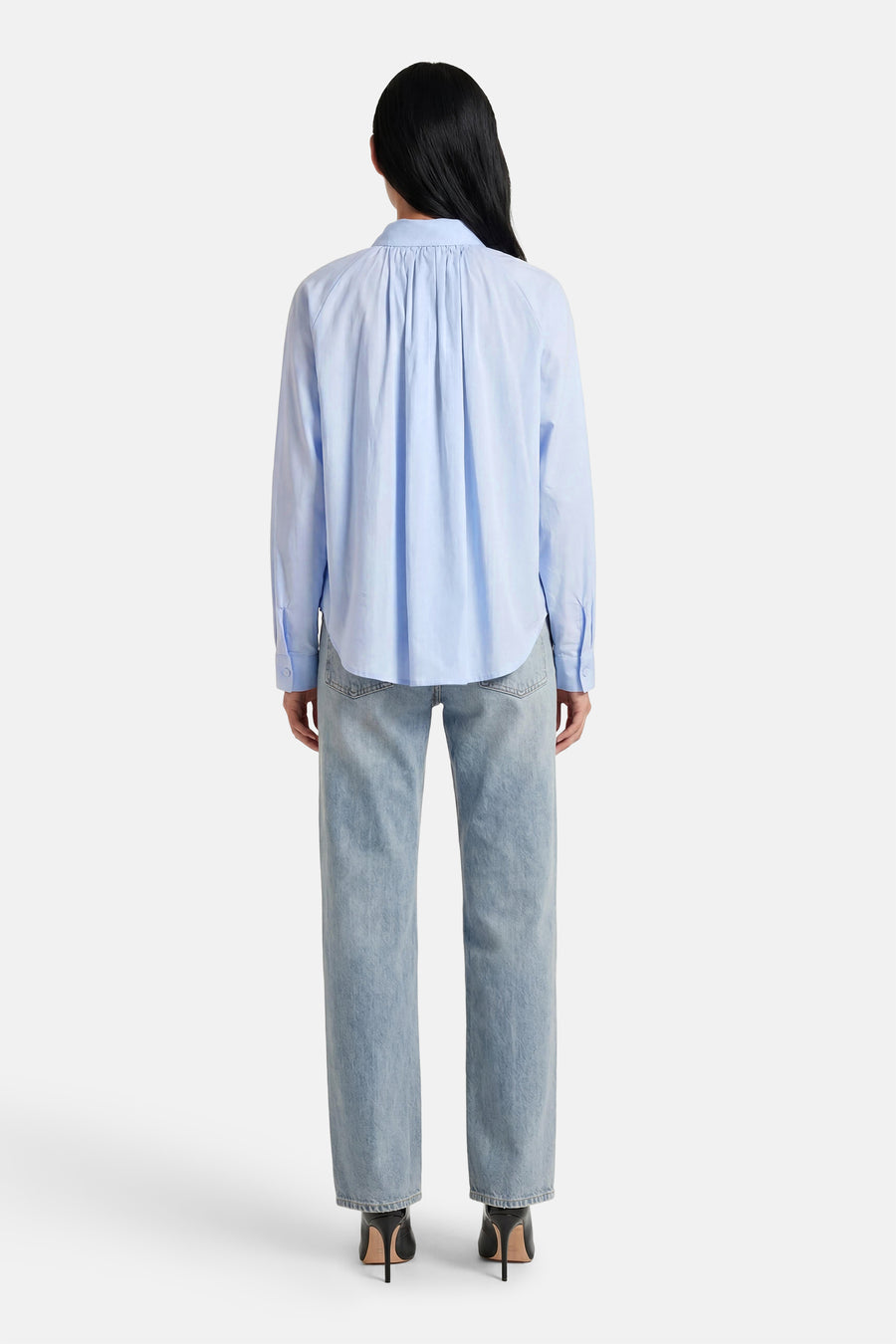Chemise bleue à manches longues de ADORE, avec détails plissés au dos, portée avec un jean et des talons noirs.
