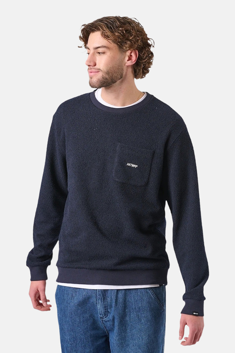 Sweat bleu van ANTWRP met poche poitrine portant le logo, porté avec un t-shirt blanc et un jean.