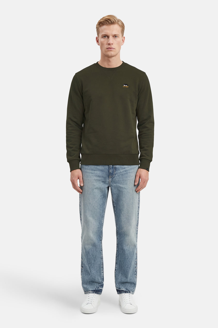 Kaki sweatshirt van ANTWRP, met geborduurd logo, gedragen met een lichtblauwe jeans en witte sneakers.