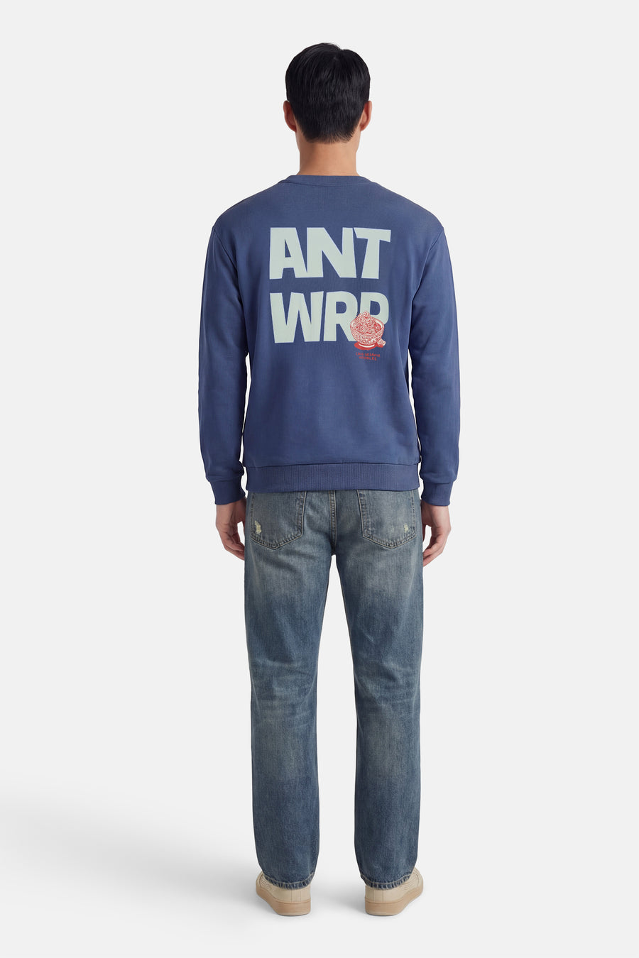 Sweat bleu ANTWRP avec inscription au dos, porté avec un jean et des chaussures couleur crème.
