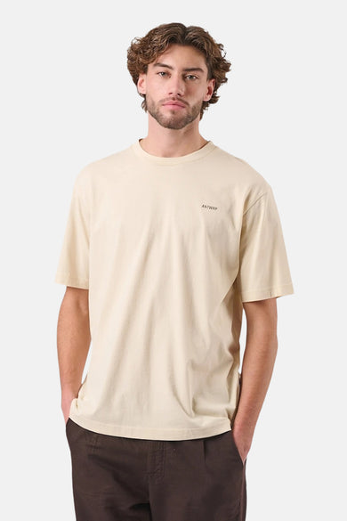 T-shirt beige à manches courtes de ANTWRP, porté avec un pantalon marron et un homme à la barbe courte.
