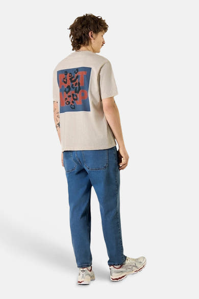 T-shirt beige ANTWRP avec motif léopard rouge et bleu sur le dos, porté avec un jean bleu et des chaussures de sport blanches.