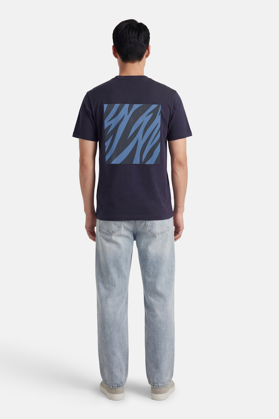 T-shirt à manches courtes bleu de ANTWRP, avec un motif graphique abstrait dans le dos, porté avec un jean clair et des chaussures beiges.
