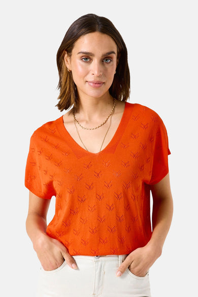 Oranje trui met V van GARCIA, met een doorzichtig patroon, gedragen met een uitgesneden witte broek en gouden platte schoenen.