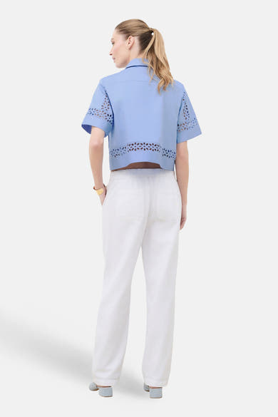 Chemise bleu clair à manches courtes de Caroll, avec découpes florales à l'ourlet et aux manches, portée avec un pantalon blanc.
