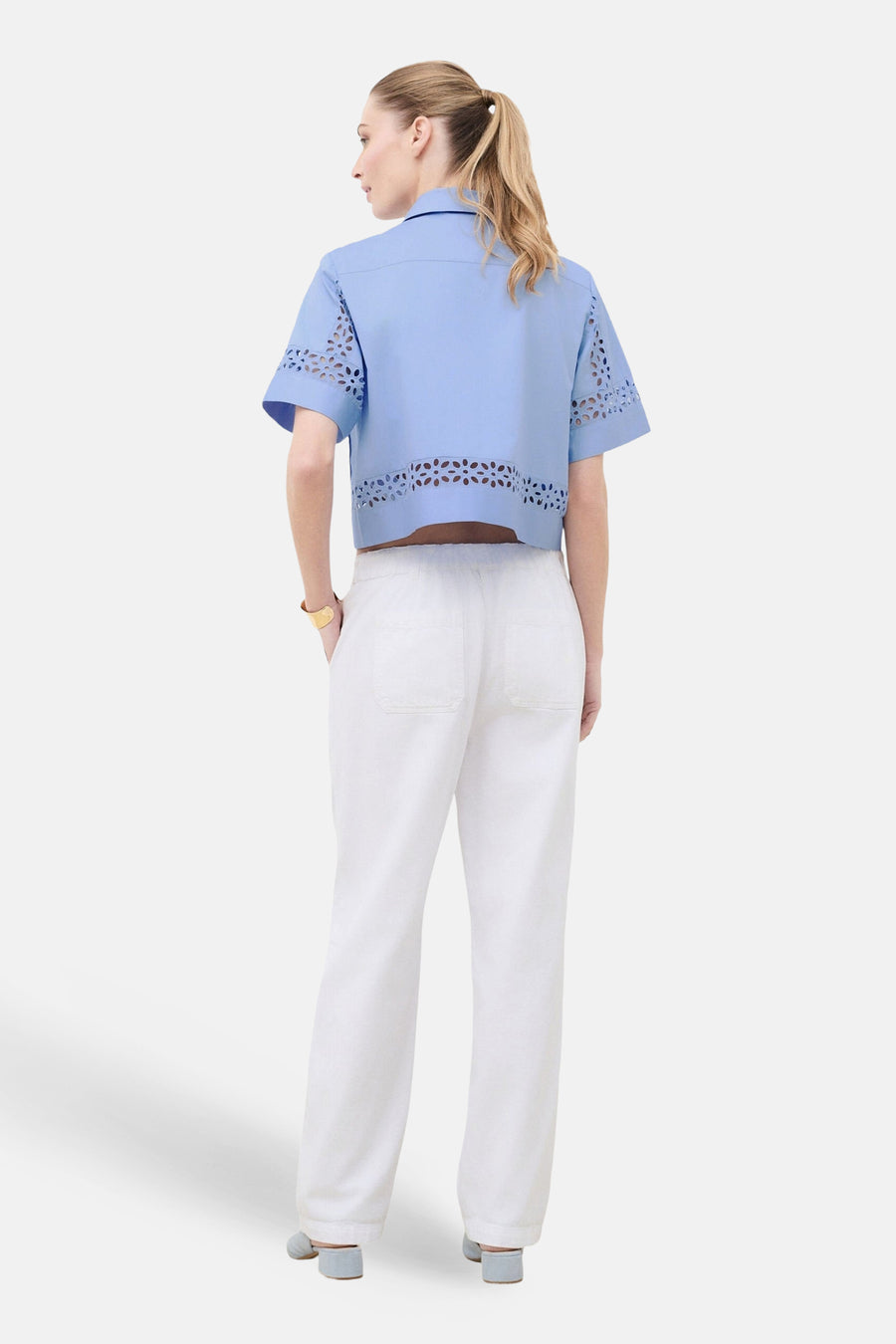Chemise bleu clair à manches courtes de Caroll, avec découpes florales à l'ourlet et aux manches, portée avec un pantalon blanc.
