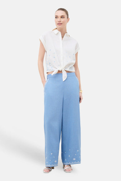 Chemise blanche à manches courtes de CAROLL avec broderies bleu clair et nœud à la taille, associée à un pantalon large bleu brodé et des sandales blanches.
