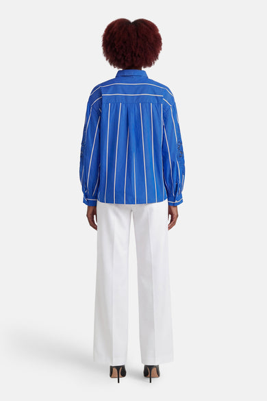 Blauwe gestreepte blouse van CAROLL, lange mouwen versierd met ajourborduursel, gedragen met een wijde witte broek en zwarte hakken.