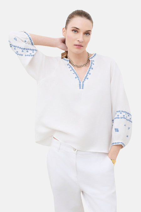 Blouse blanche brodée de CAROLL à encolure en V et motifs bleus sur les manches, portée avec un pantalon blanc et un collier doré.