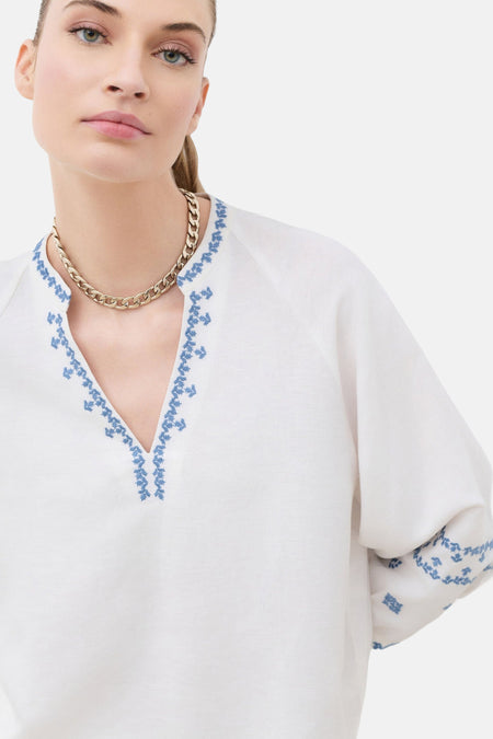 Blouse blanche en lin de CAROLL à col en V brodé de motifs bleus, portée avec un collier chaîne doré.
