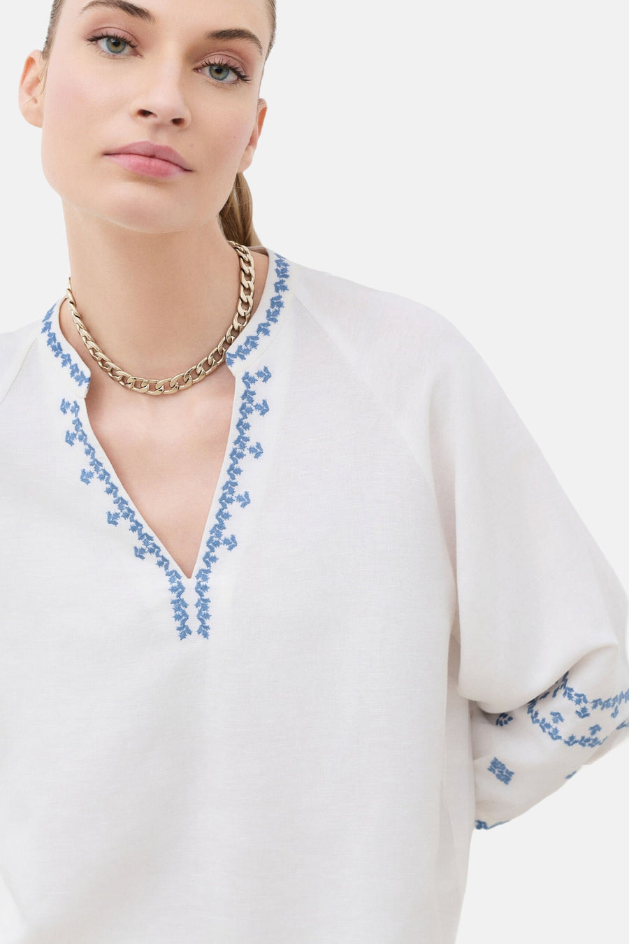 Blouse blanche en lin de CAROLL à col en V brodé de motifs bleus, portée avec un collier chaîne doré.