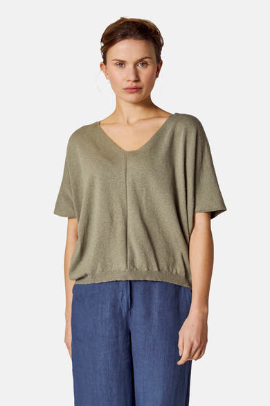 Top vert en tricot de COPINES D'ANVERS, avec un col en V et des manches courtes, porté avec un pantalon bleu.
