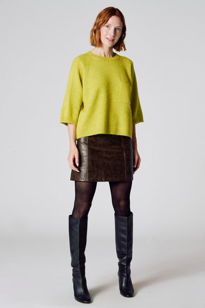 Groene trui met ronde hals van COPINES D'ANVERS, driekwartmouwen, gedragen met een bruine leren rok en zwarte laarzen.