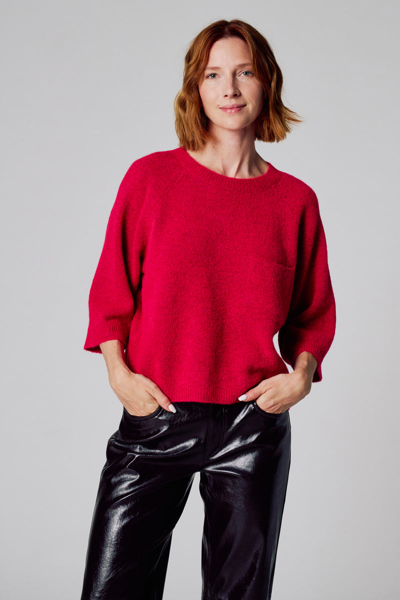 Pull rose à col rond de COPINES D'ANVERS, porté avec un pantalon en vinyle noir brillant.