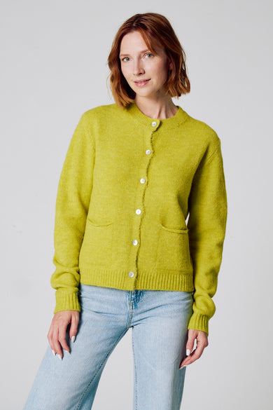Groene cardigan van COPINES D'ANVERS, met witte knopen, gedragen over een lichtblauwe jeans.
