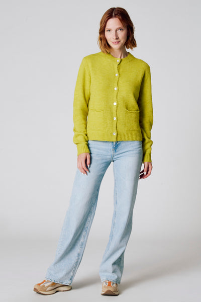 Groene cardigan van Copines d'Anvers, met witte knopen, gedragen met een lichte jeans en sneakers.