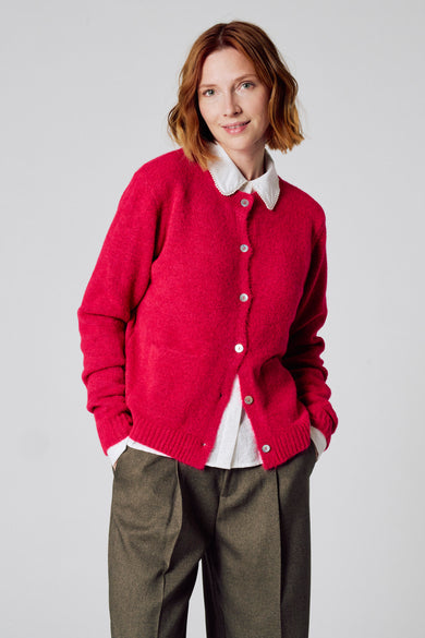 Cardigan rose de COPINES D'ANVERS porté sur une chemise blanche à col festonné, avec un pantalon marron à plis.

